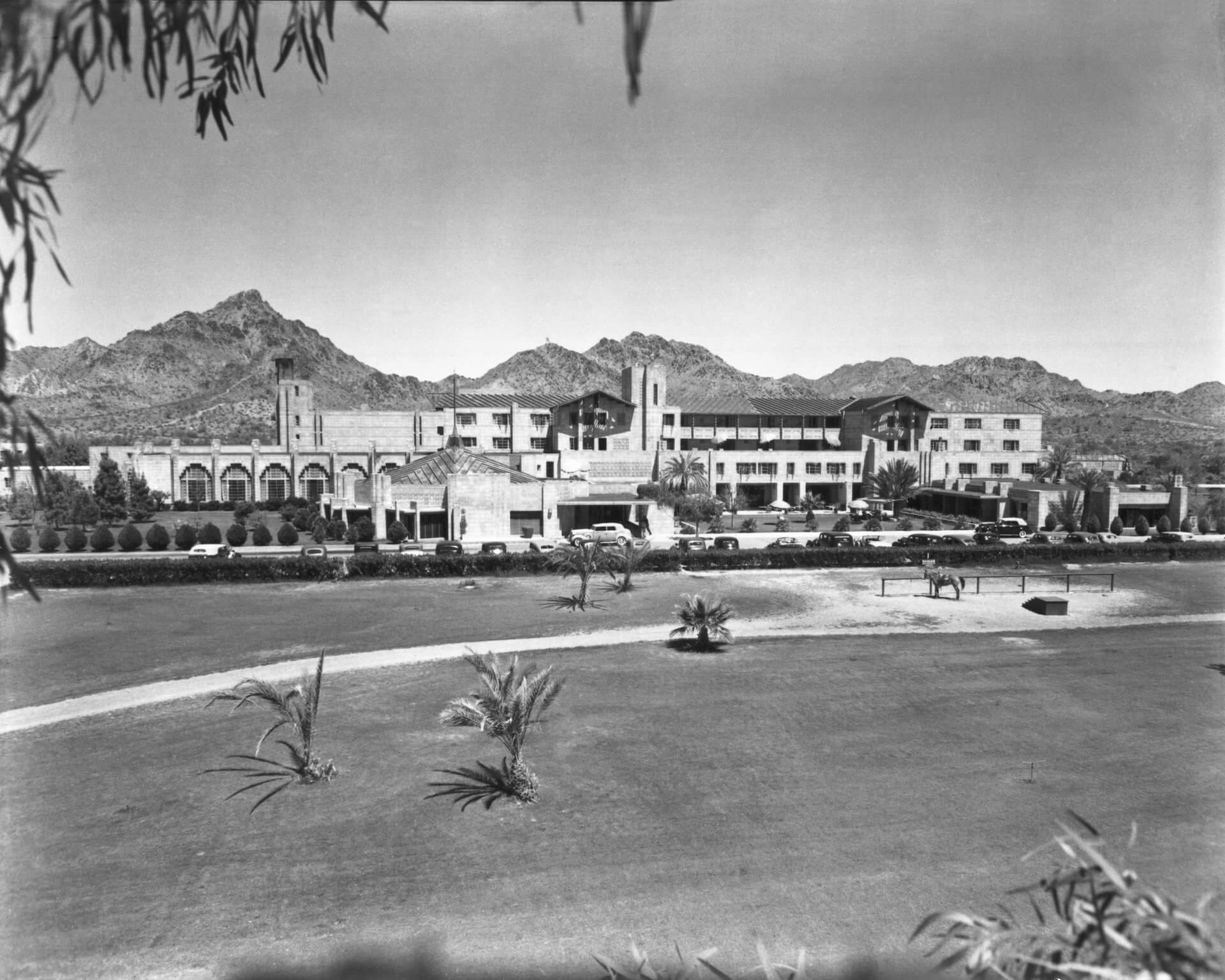 Black and white photo of a large resort-style building with Spanish architectural elements set against a backdrop of distant mountains. In the foreground, about an expanse of neatly manicured lawns, stand a few scattered palm trees.
