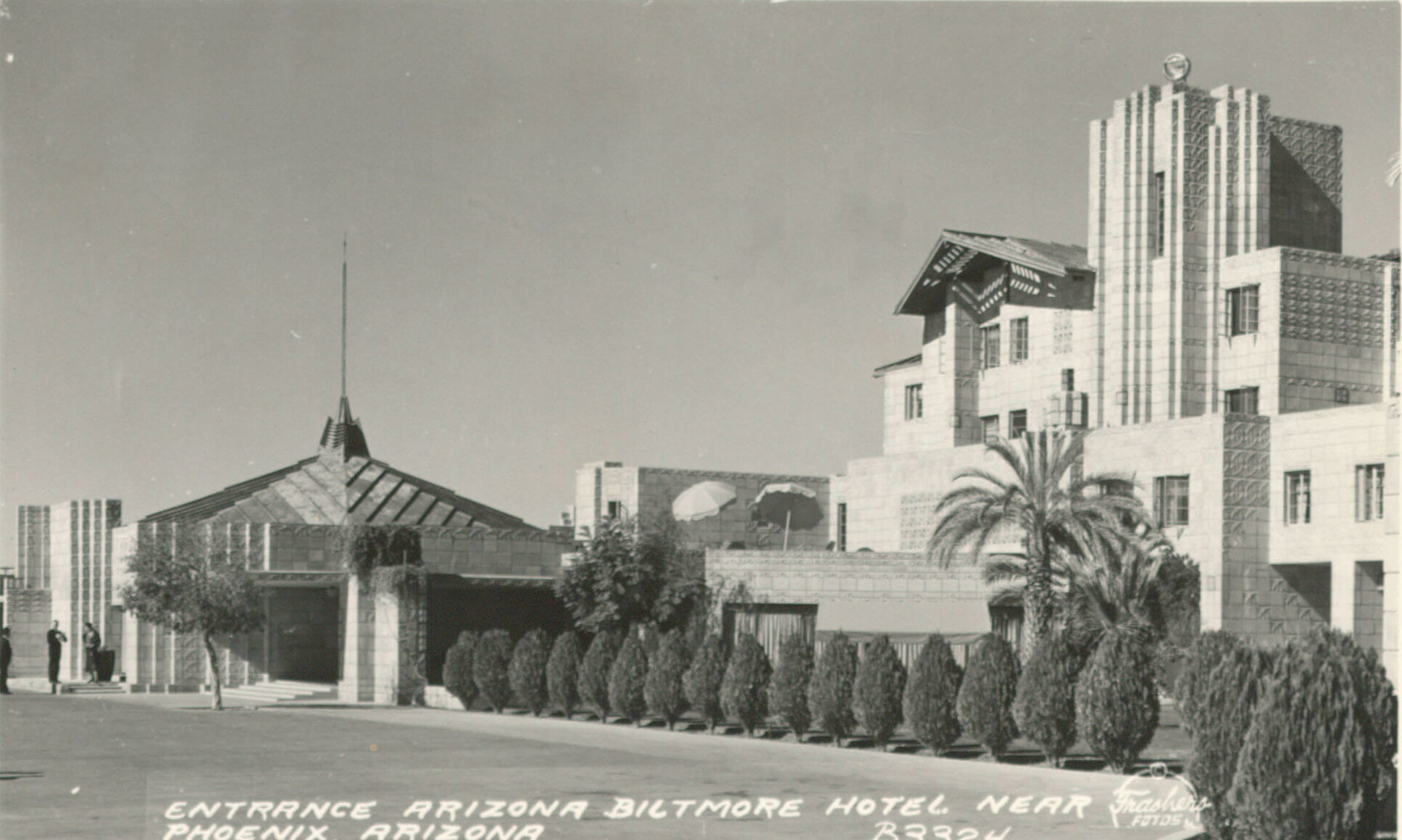Black and white photograph showing the entrance of the Arizona Biltmore Hotel near Phoenix, Arizona. The hotel features distinctive architecture with geometric Biltmore Block designs and several lush palm trees leading up to the entrance. Text at the bottom reads "ENTRANCE ARIZONA BILTMORE HOTEL NEAR PHOENIX ARIZONA.