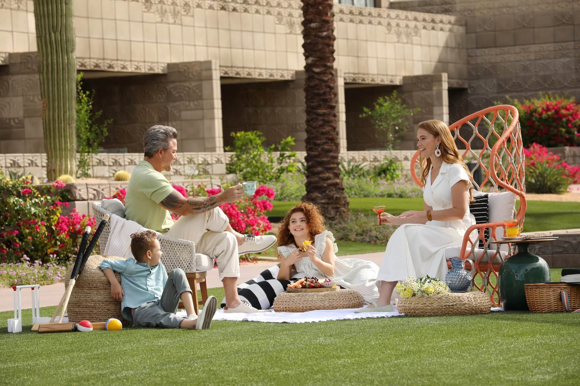 A family of four enjoys a picnic on a lush green lawn. The adults sit on chairs while the children sit on a blanket. They are surrounded by vibrant flowers and greenery, with a tall cactus and a stone building in the background—an idyllic scene for those seeking relaxing things to do outdoors. Everyone looks relaxed and happy.
