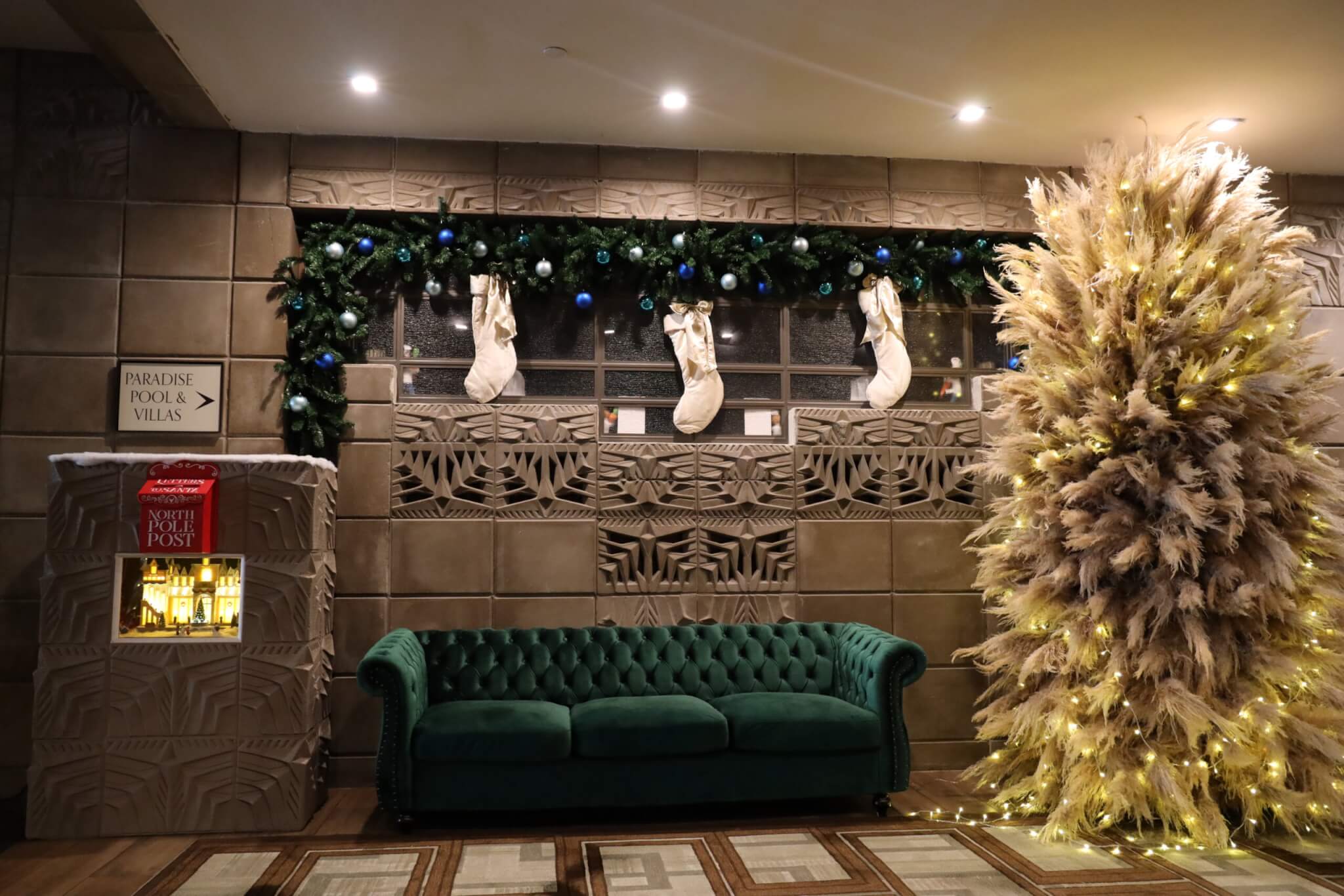 A festive lobby features a green velvet couch, a pampas grass Christmas tree with yellow lights, three stockings hanging above, and a garland with blue ornaments. A small "North Pole Post" box is on the left wall.