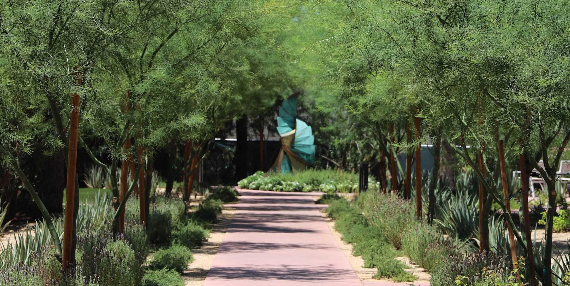 A serene garden path lined with lush green trees leads to a vibrant abstract sculpture by Heloise Swaback Crista. The pathway is bordered by various shrubs and small plants, providing a tranquil, natural setting.