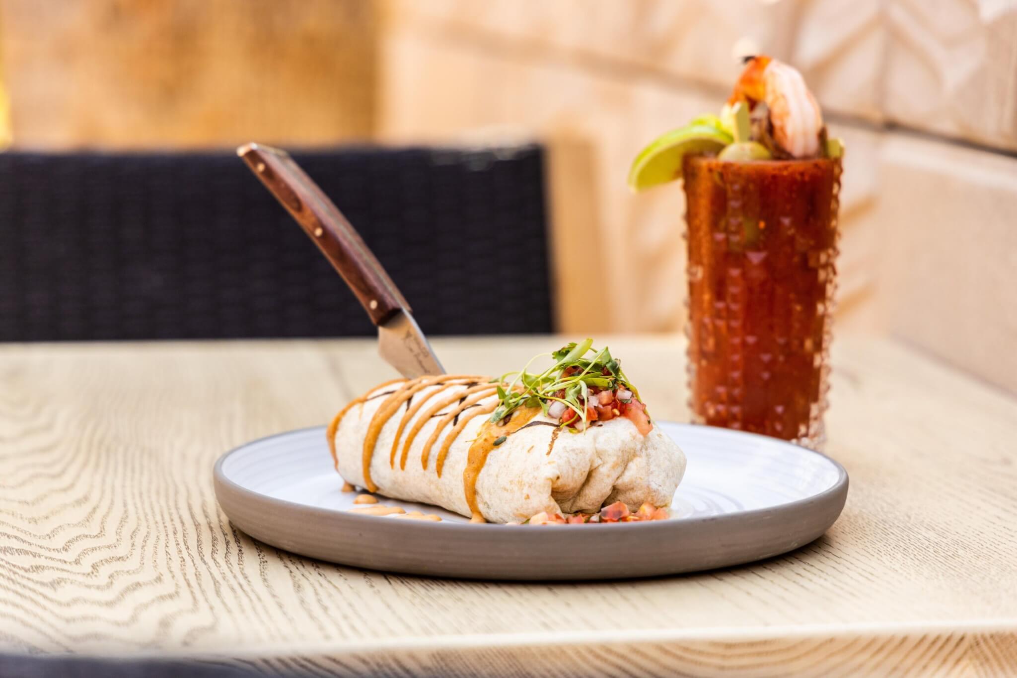A burrito topped with sauce and microgreens sits on a plate with a knife stuck in it, next to a glass of a cocktail garnished with shrimp, lime, and celery, on a wooden table.