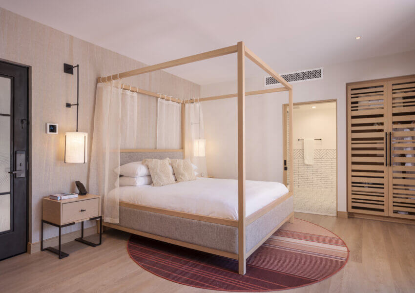 A cozy bedroom in the cottage suite features a modern four-poster bed with white bedding and sheer drapes. The room has wooden flooring, a round red carpet under the bed, a nightstand with a lamp, and an adjoining bathroom with white tiles. Soft light creates a warm ambiance.