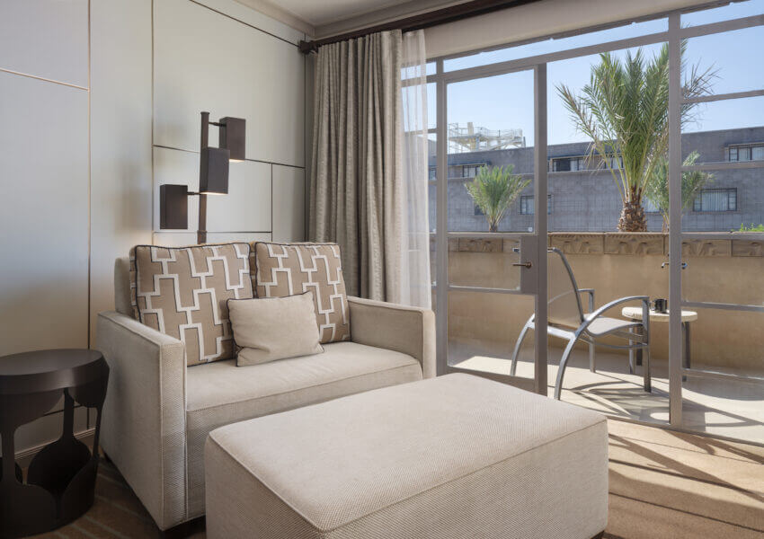 A modern hotel room features a beige sofa with patterned cushions, a matching ottoman, and a sleek black side table. The room is well-lit with floor-to-ceiling windows that lead to a balcony with a view of palm trees and adjacent buildings.