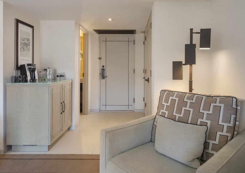 A modern hotel room entrance with a beige armchair featuring geometric patterns in the foreground. To the left, there's a wooden cabinet with a coffee maker and cups. The entrance door, lit hallway, and a partially visible bathroom door are in the background.