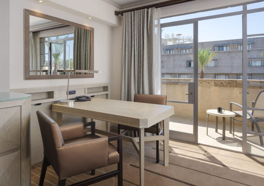 A modern hotel room featuring a desk with two brown leather chairs, a mirror on the wall, and a glass door leading to a small balcony. The balcony has a small table and chairs, overlooking neighboring buildings and palm trees under clear skies.