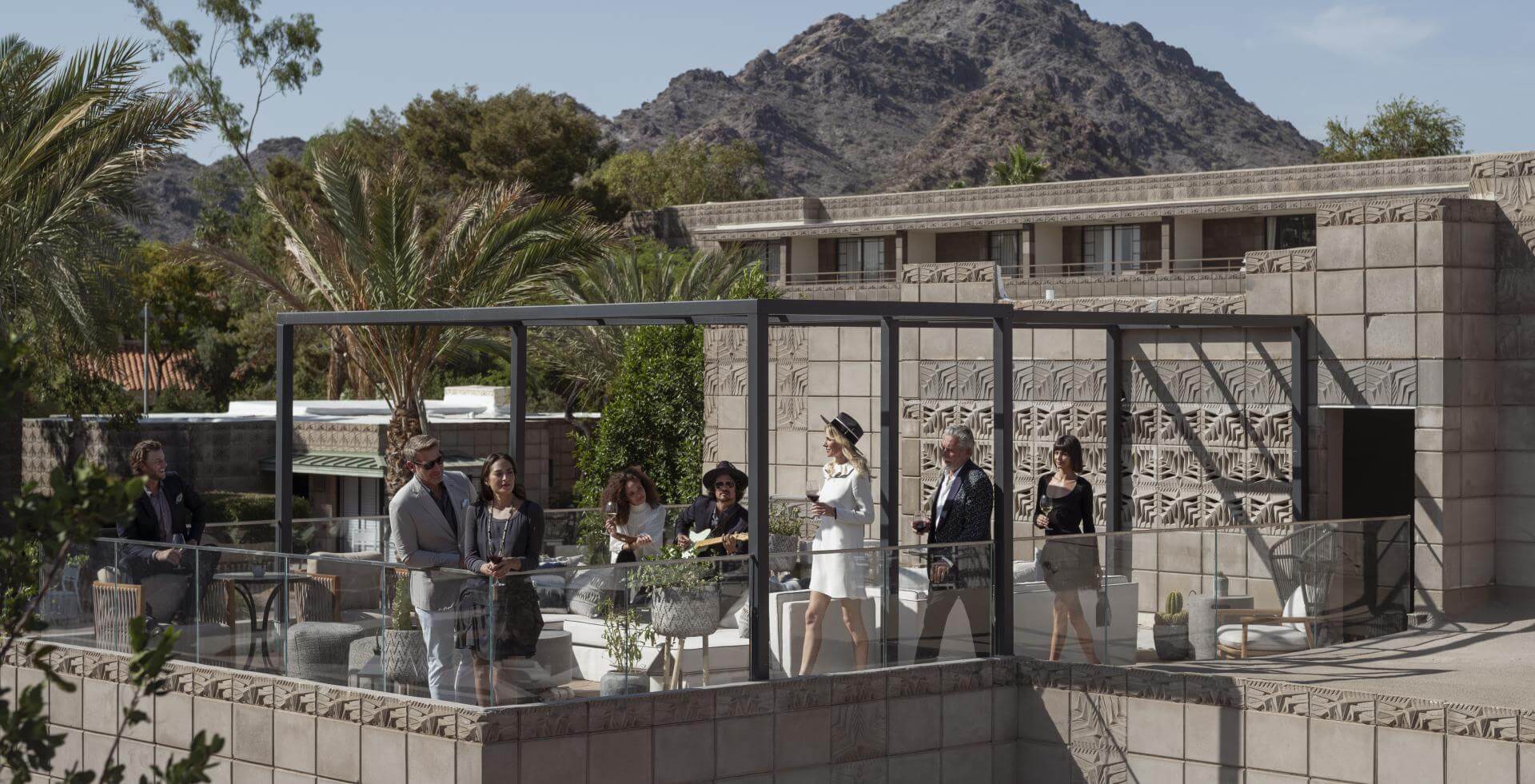 A group of people gathers on a modern terrace with large windows and elegant decor. Some are seated while others stand and converse, reminiscent of informal meetings. The terrace is surrounded by trees and mountains under a clear sky, with a building featuring geometric patterns in the background.