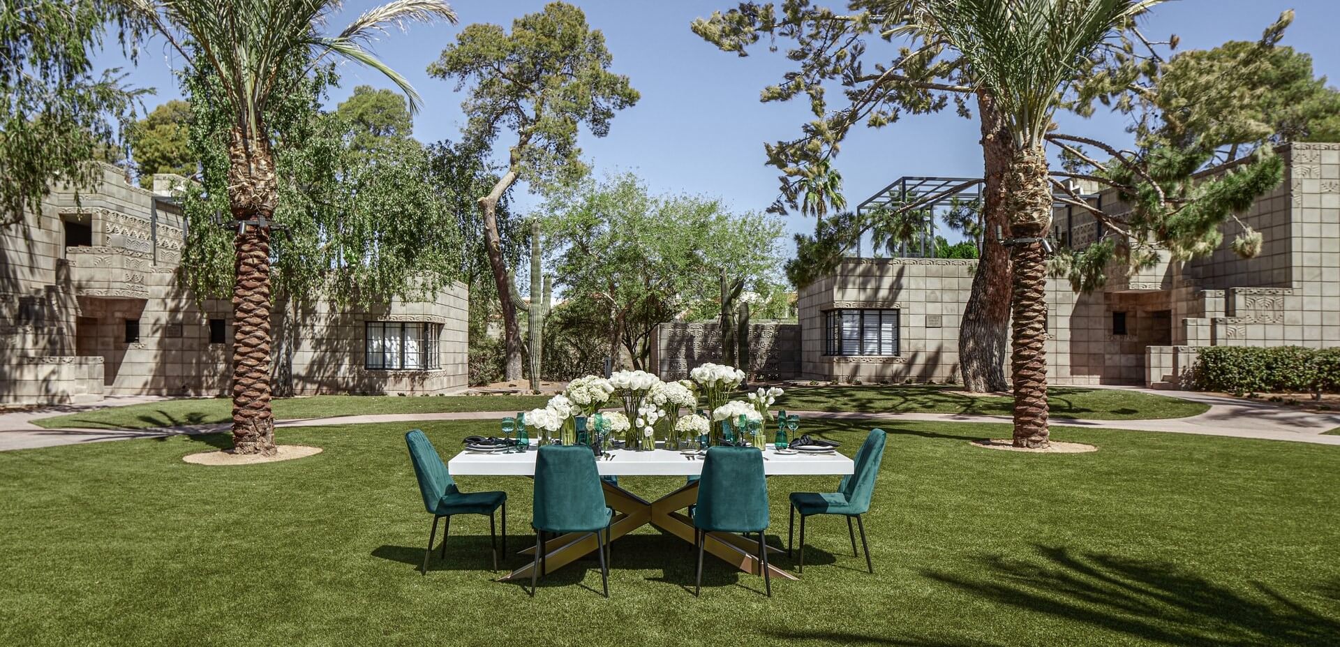 An outdoor dining table with a white tablecloth and teal chairs is set on a well-manicured lawn, surrounded by tall palm trees and lush greenery. Stone buildings with geometric patterns are in the background under a clear blue sky.