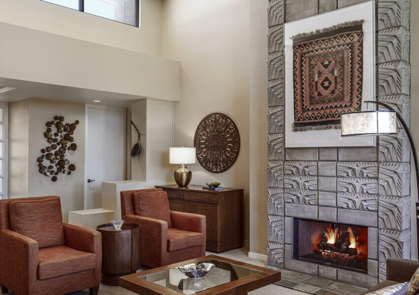 A cozy living room featuring a modern fireplace with a large, decorative tapestry above it. Two rust-colored armchairs are positioned near a wooden coffee table. The room includes a sideboard with a lamp, wall art, and natural light from high windows.