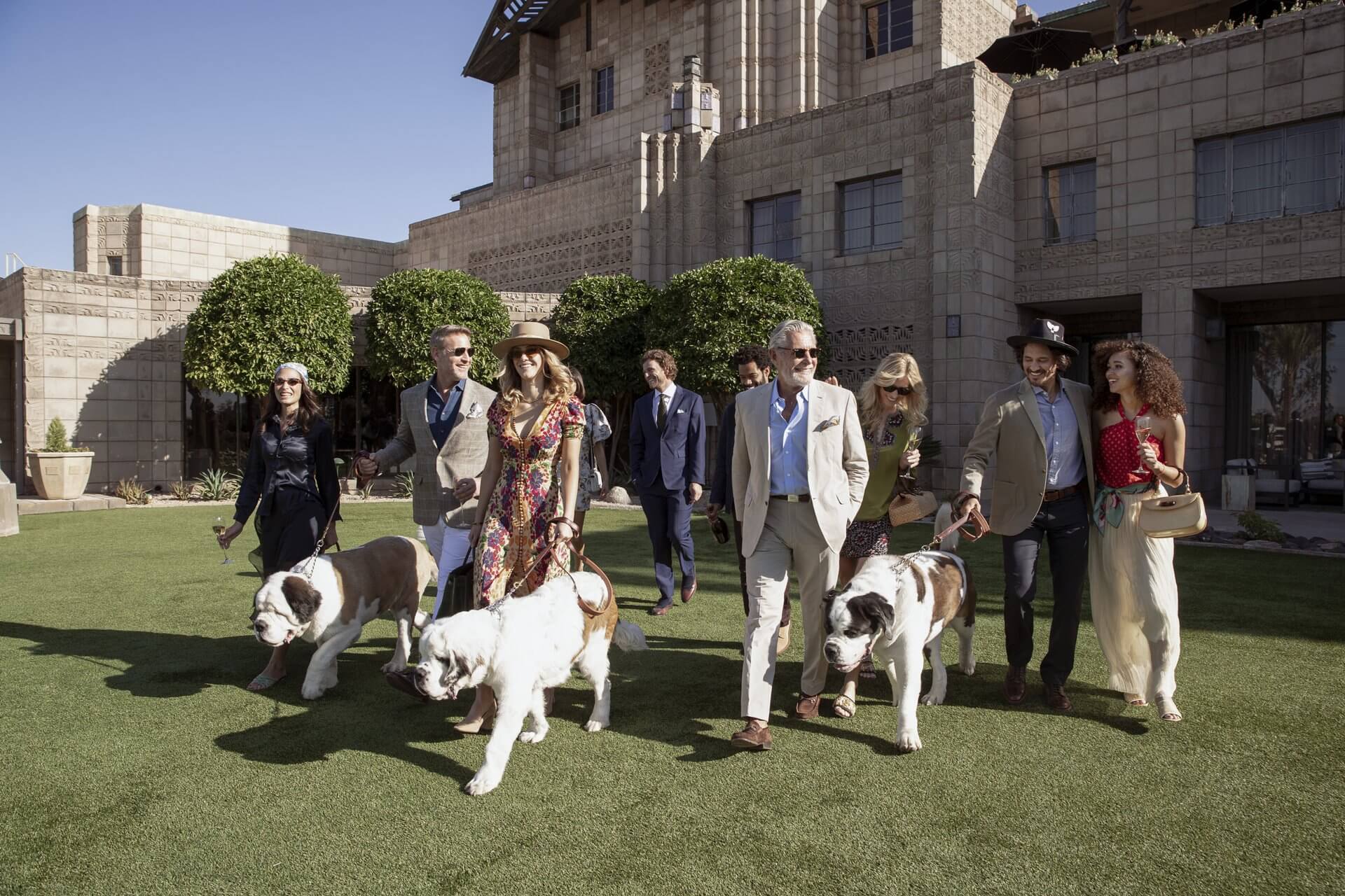 A group of well-dressed people walk on a lawn in front of a large, elegant building, resembling a high-end retail ad. They are accompanied by three Saint Bernard dogs on leashes. The diverse group is wearing formal and semi-formal attire, with some sporting hats and sunglasses under the clear blue sky.