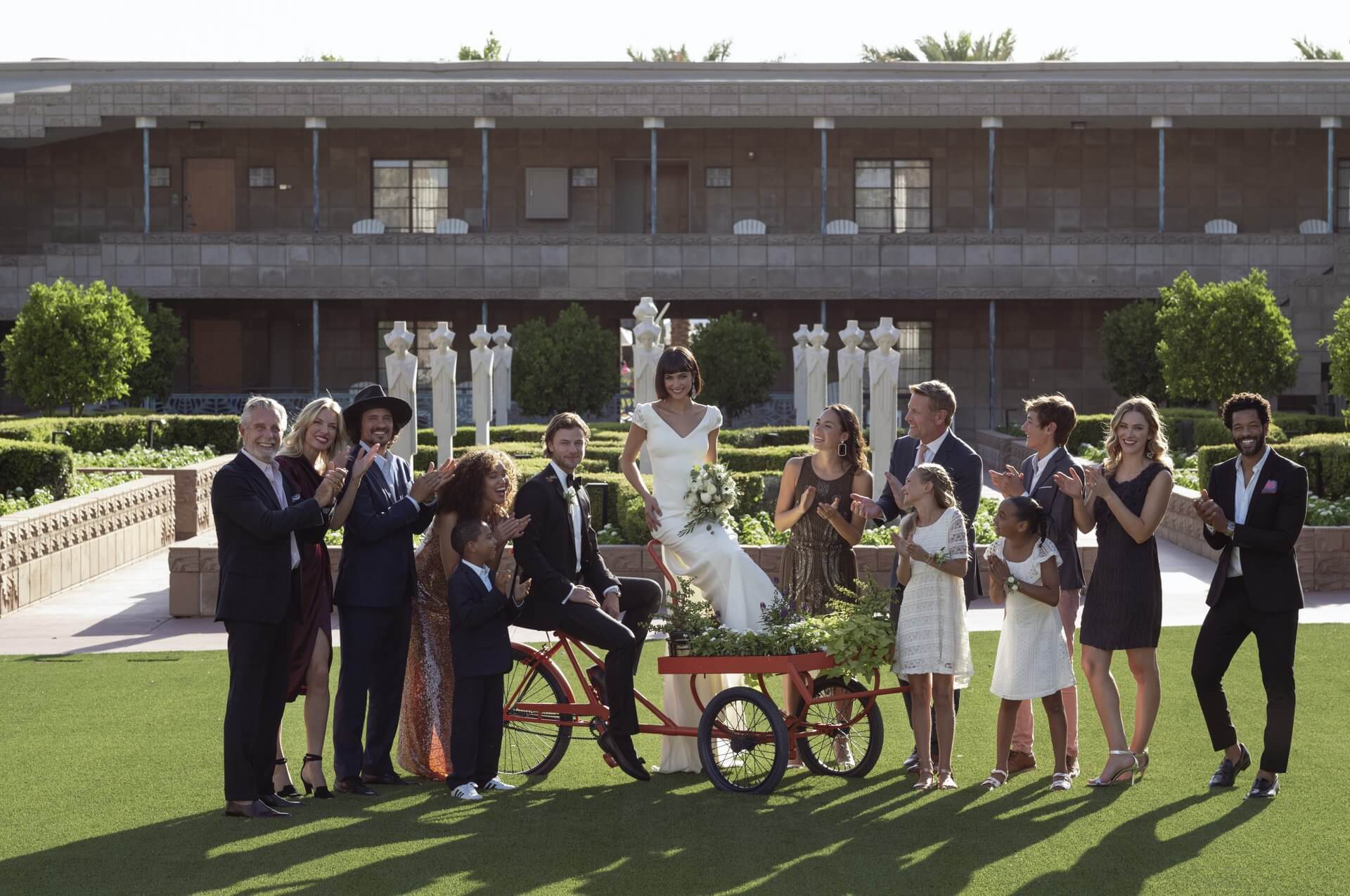 A group of people surround a couple standing on a tricycle cart with plants, in front of a building. The woman in a white dress is elevated, holding flowers, while the rest, elegantly dressed, applaud and smile on a lush green lawn.