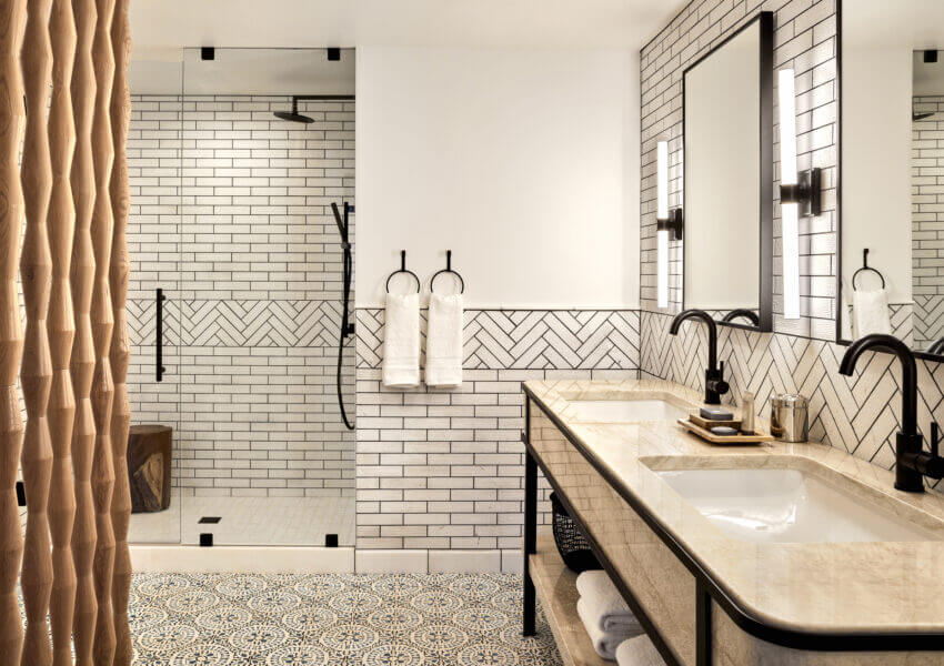 A modern cottage suite bathroom features a walk-in glass shower with a black showerhead and white subway tile. The vanity has a double sink with black fixtures and a large mirror. White towels hang next to the sinks, and the floor is adorned with intricate patterned tiles.