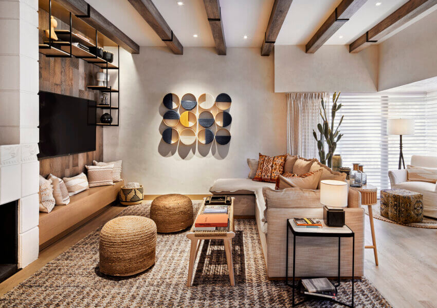 A cozy living room featuring a beige sectional sofa with various patterned pillows, wicker ottomans, a textured gray rug, and geometric wall art as the focal point. This cottage suite also boasts wooden ceiling beams, a large flat-screen TV, and abundant natural light.