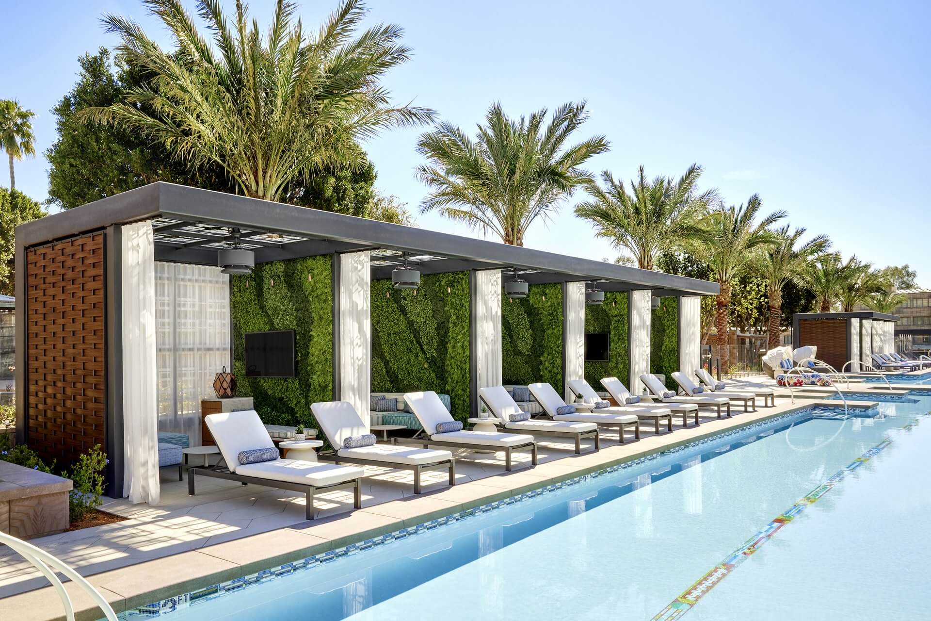 A row of luxurious poolside cabanas with lounge chairs, small tables, and plush cushions, embodying a reinvention of relaxation, are lined up along a sparkling swimming pool. Palm trees and greenery provide a tropical ambiance under a clear blue sky.