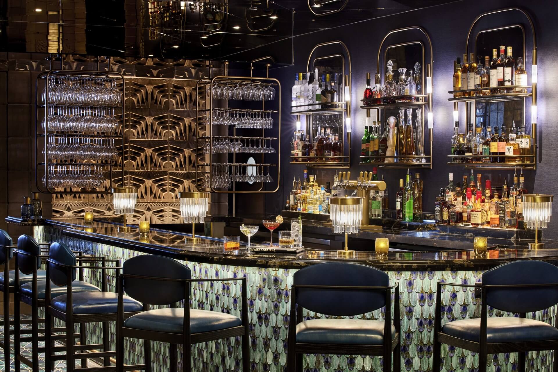 A modern upscale bar with an illuminated counter and black chairs invites you to dine in style. Glasses hang overhead, and colorful bottles line the shelves. Fancy light fixtures and intricate wall patterns enhance the elegant ambiance. A few drinks are placed on the bar.