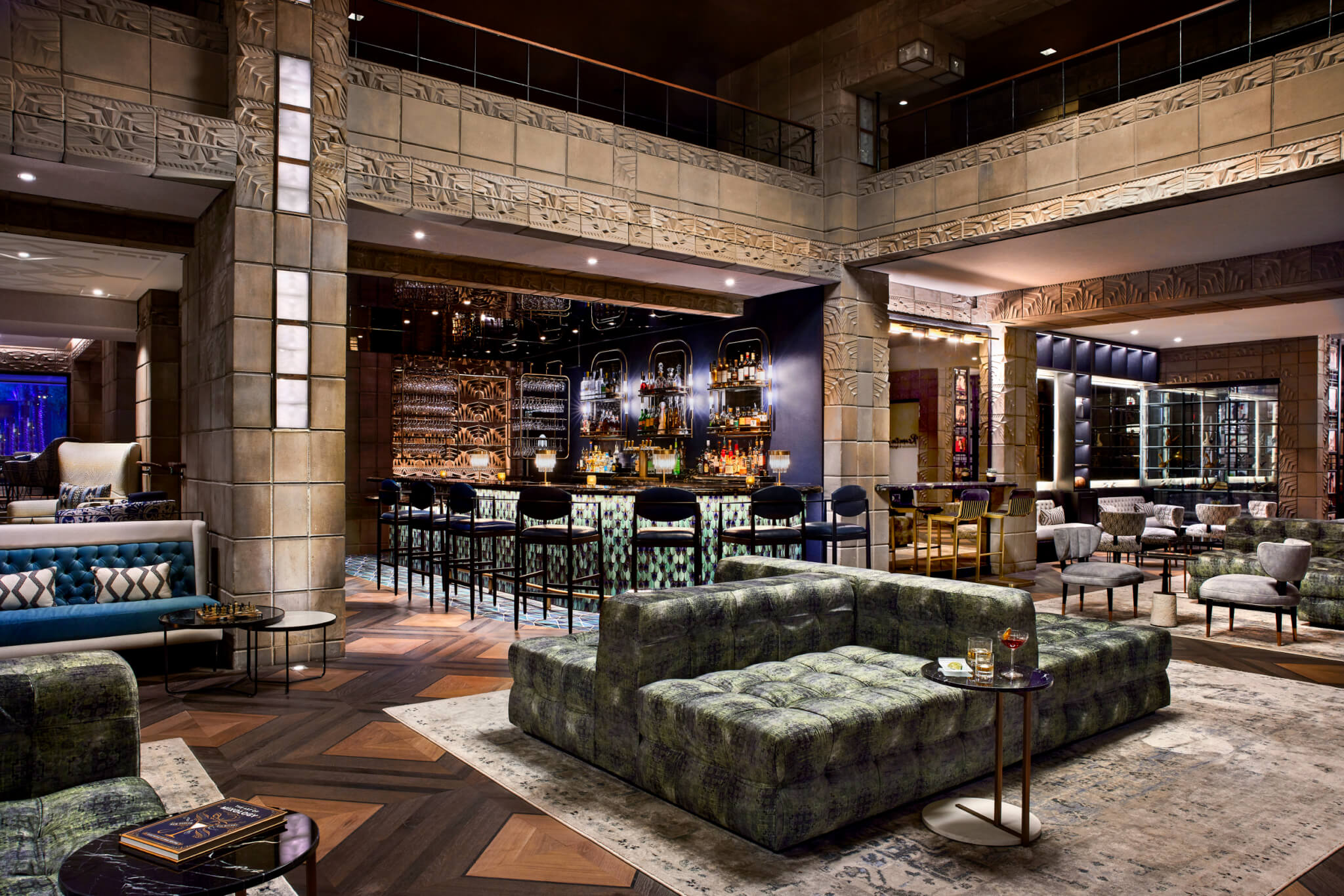 A stylish, modern lounge area featuring a central Wright bar with high stools, surrounded by cushioned seating, including sofas and armchairs. The space is elegantly decorated with intricate architectural details, warm lighting, and a mix of contemporary and rustic furnishings.