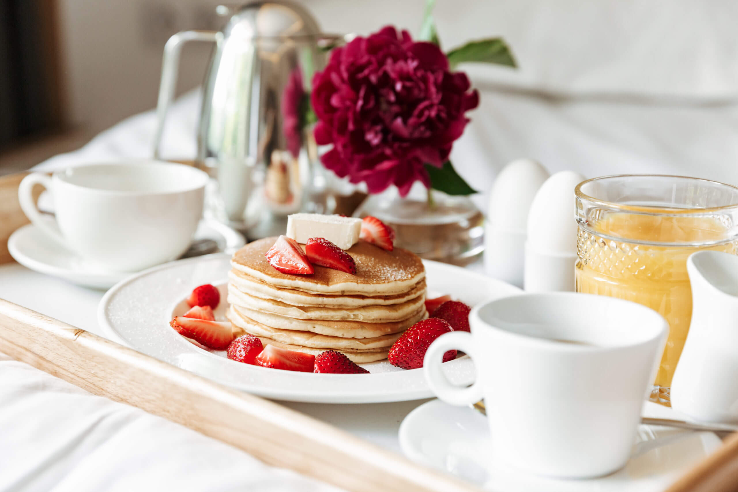 A breakfast tray on a bed features a stack of pancakes topped with butter and sliced strawberries, a glass of orange juice, a coffee pot, white cups, a boiled egg in an egg cup, and a vibrant red flower in a vase.