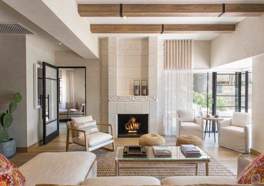 A cozy living room invites you to stay with its lit fireplace framed by white tiles. The room features a beige couch adorned with colorful pillows, wooden chairs, and a glass coffee table. Brightened by floor-to-ceiling windows and light curtains, the space also boasts exposed wooden ceiling beams above.