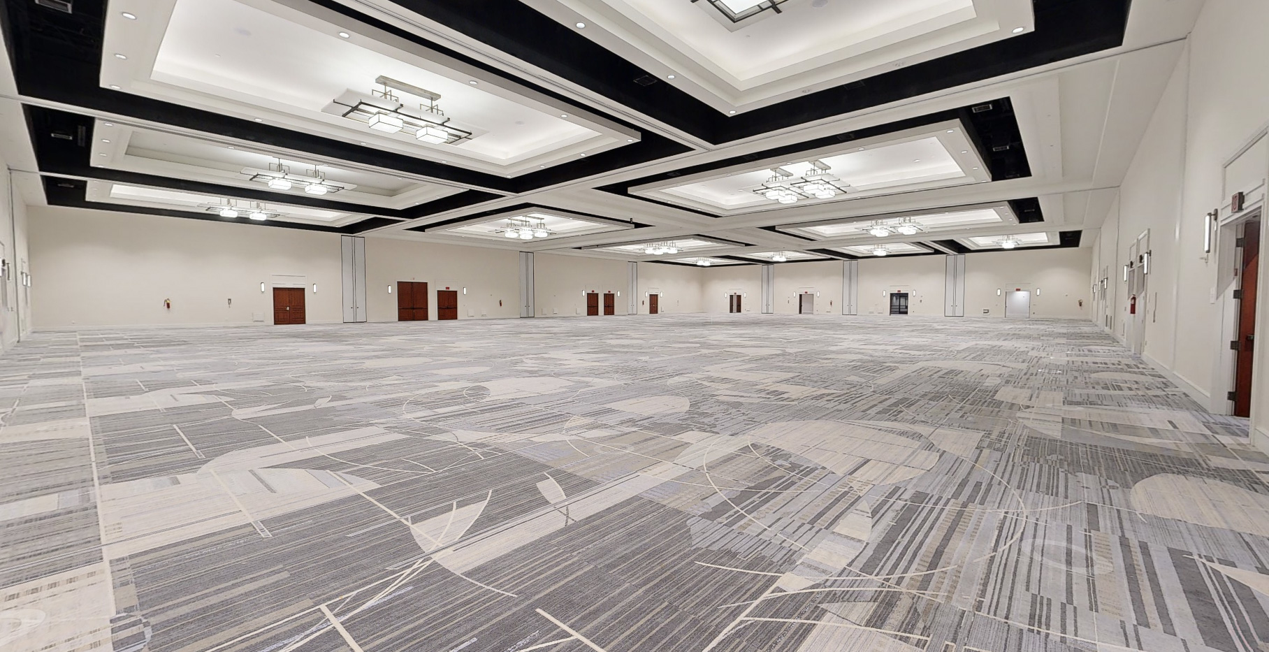 A large, empty convention hall with patterned gray carpet, high white ceilings, modern light fixtures, and multiple wooden doors along the walls.
