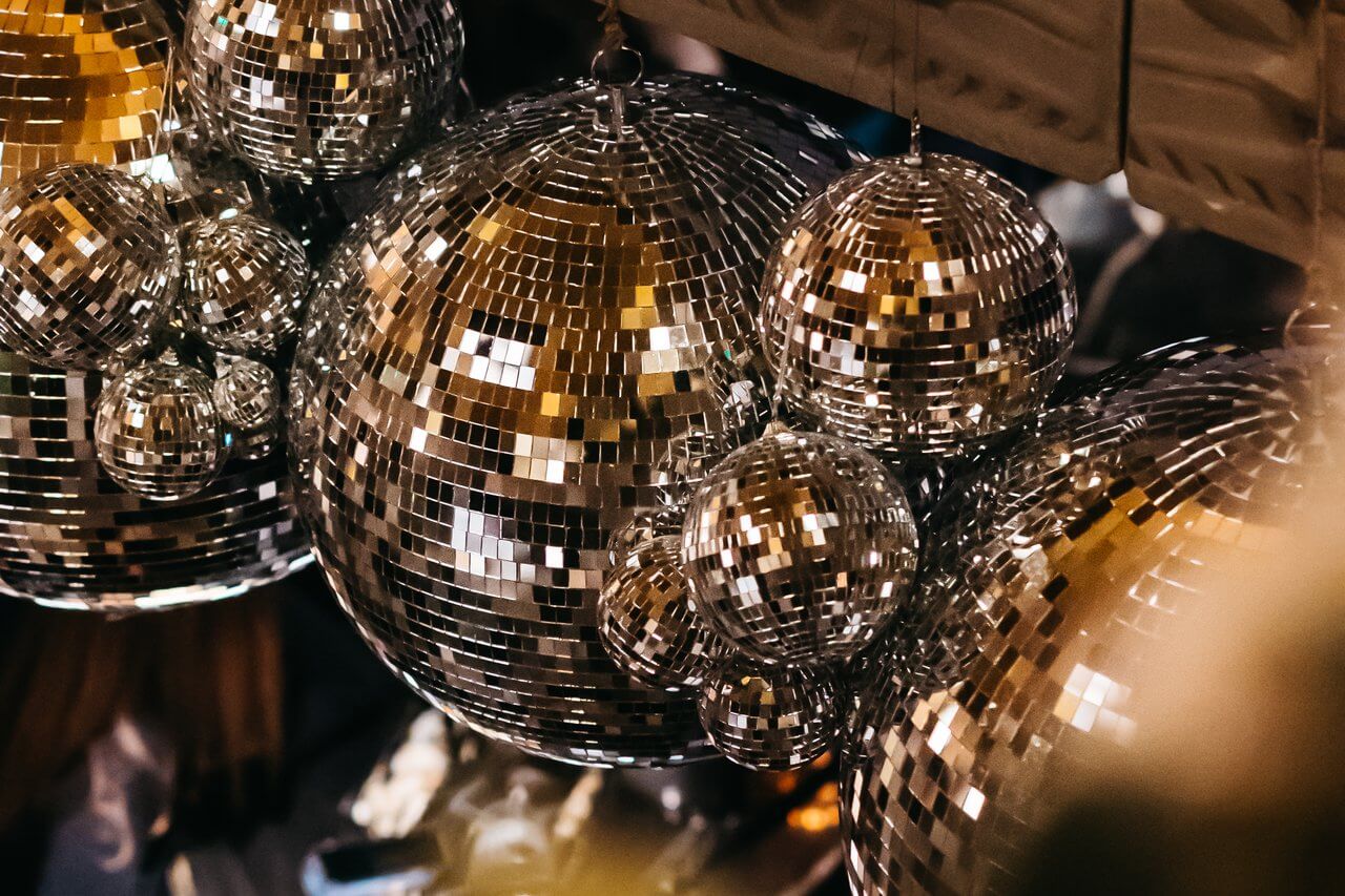 A close-up of several reflective disco balls of varying sizes hanging together creates a dazzling display of light and reflections in the dimly lit room. The mirrored surfaces catch and scatter light, enhancing the festive atmosphere—perfect for a sparkling New Year's Eve celebration.