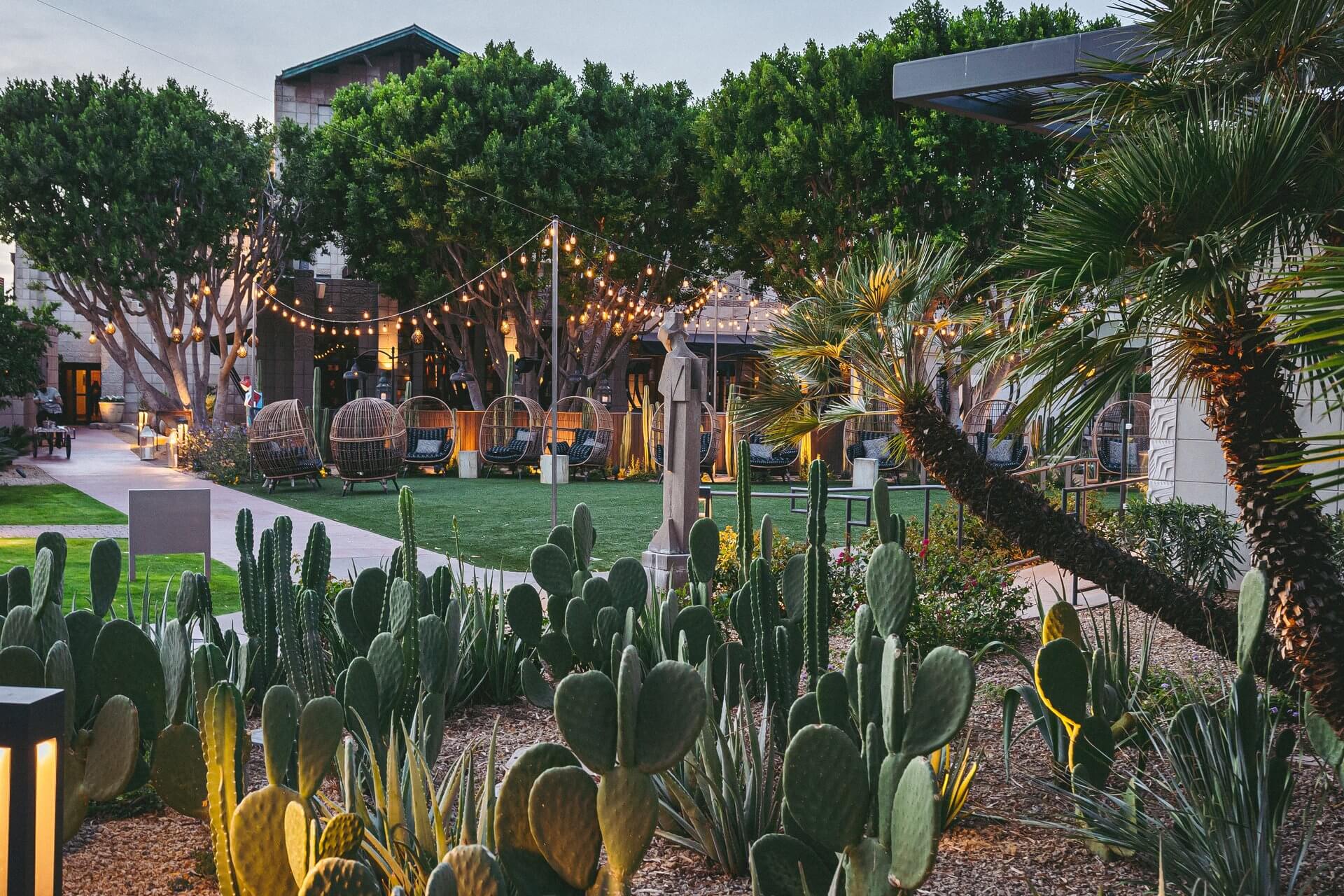 A charming outdoor setting at the Arizona Biltmore features a lush lawn surrounded by various plants, including succulents, cacti, and palm trees. String lights hang above rows of hanging egg chairs and outdoor seating, creating a cozy ambiance for guests at dusk under large, leafy trees.