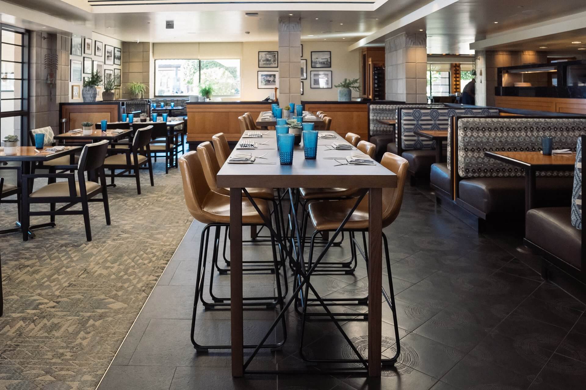 A modern restaurant interior with a mix of high and low tables, wooden chairs, and patterned cushioned booths reminiscent of a paradise garden. Blue glasses and silverware are neatly arranged on the tables. Walls are adorned with framed pictures, and windows let in natural light.