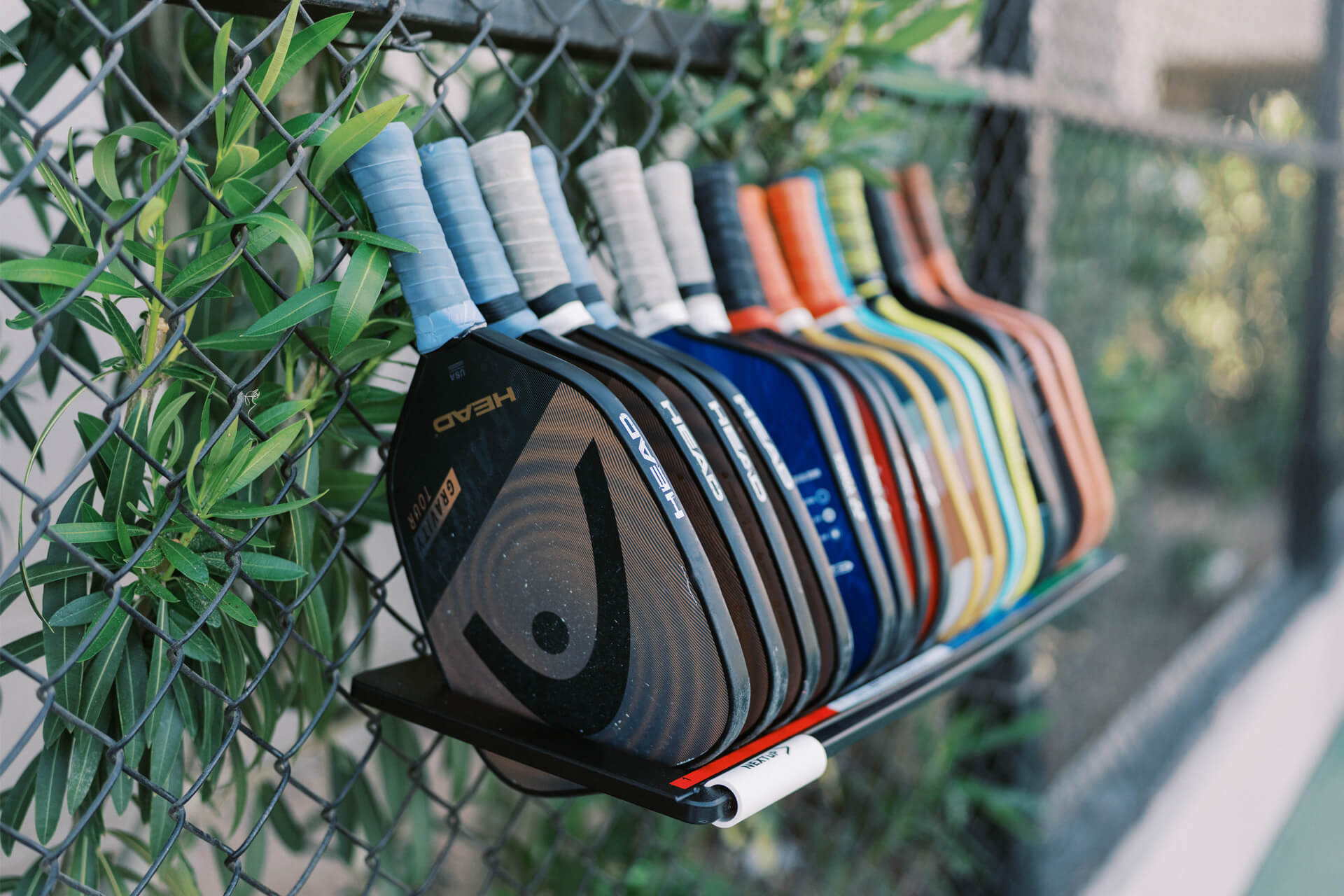 A row of assorted pickleball paddles with taped handles hangs on a black metal rack attached to a chain-link fence, celebrating National Pickleball Month, with green leaves visible in the background.