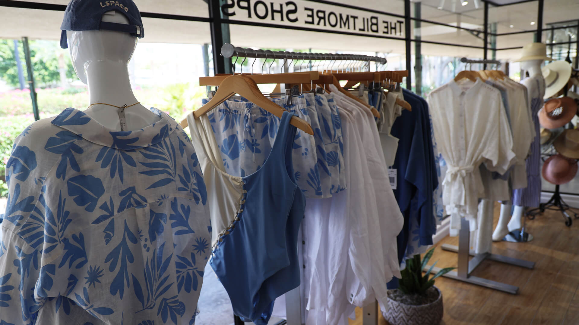 A retail display of summer clothing, including blue and white swimsuits, dresses, and cover-ups, hangs on racks inside a boutique with large windows and natural light. A mannequin in a printed blue and white shirt stands in the foreground.