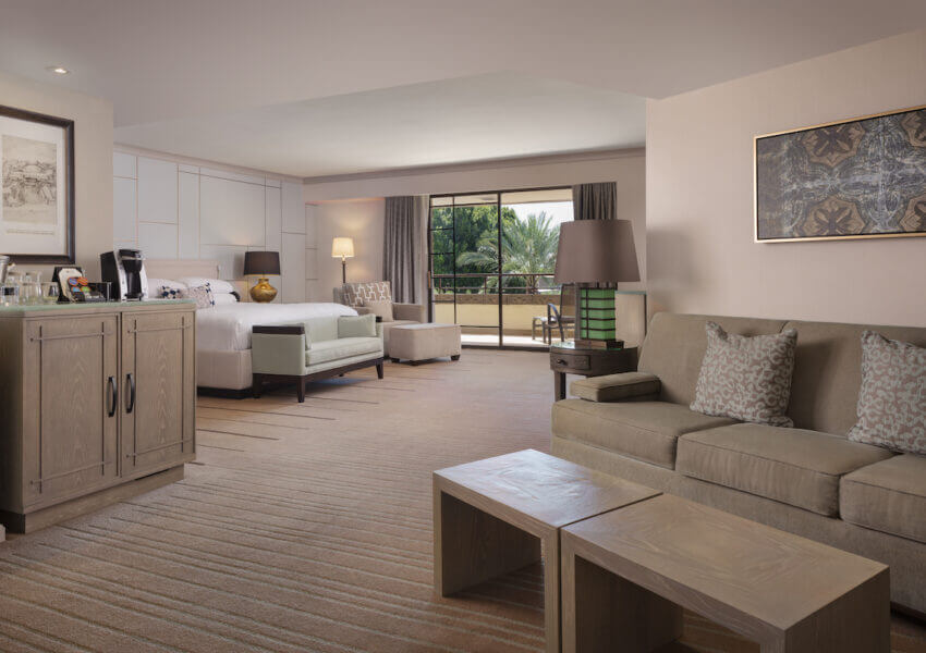 Spacious junior suite with a king-sized bed, a sofa set, a coffee station atop a cabinet, and a small wooden table. The room features a flat-screen TV, artwork on the walls, and a large sliding glass door leading to a balcony with views of greenery.