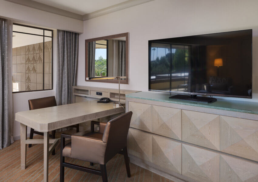 A junior suite with modern furnishings, featuring a wooden desk with a brown leather chair, a flat-screen TV on top of a geometric-patterned cabinet, tall gray curtains, a large wall mirror, and a window reflecting outdoor greenery. A phone sits on the desk.