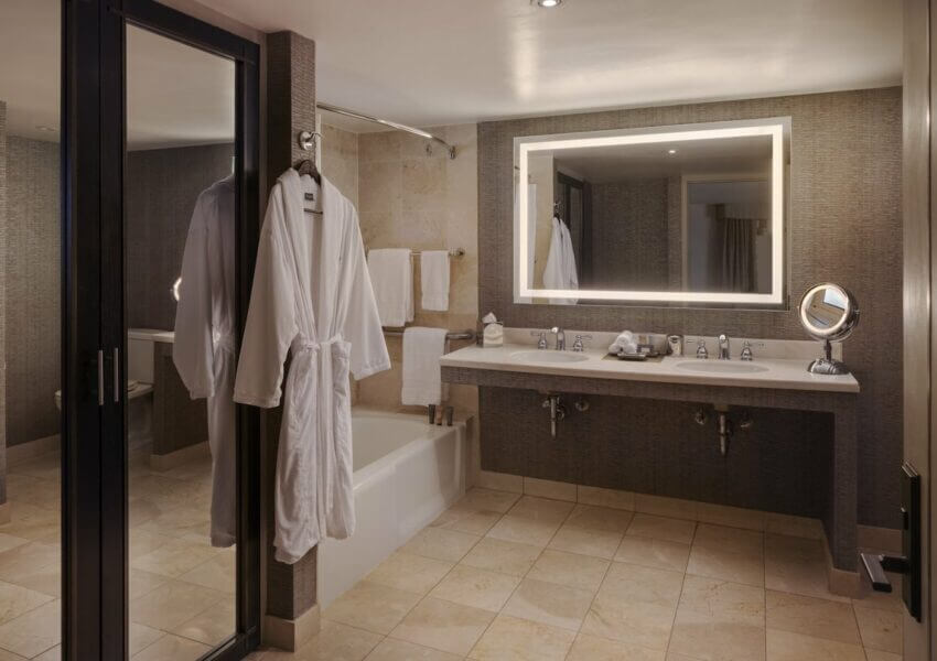 A modern junior suite bathroom featuring a bathtub with hanging white robes, a large mirror with backlighting above a double-sink vanity, and neatly arranged towels. The room is decorated with light beige and grey tones, creating a clean and elegant atmosphere.