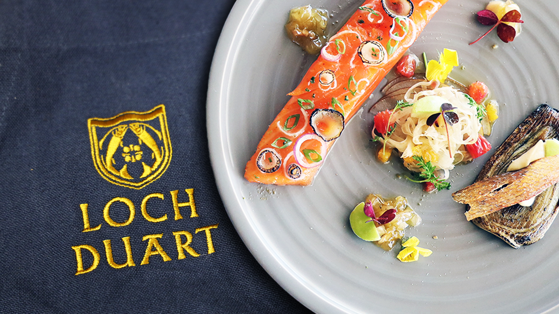 A gourmet dish featuring a fillet of salmon garnished with herbs and vegetables, artfully plated on a gray dish next to a Loch Duart napkin with a yellow embroidered logo and text.