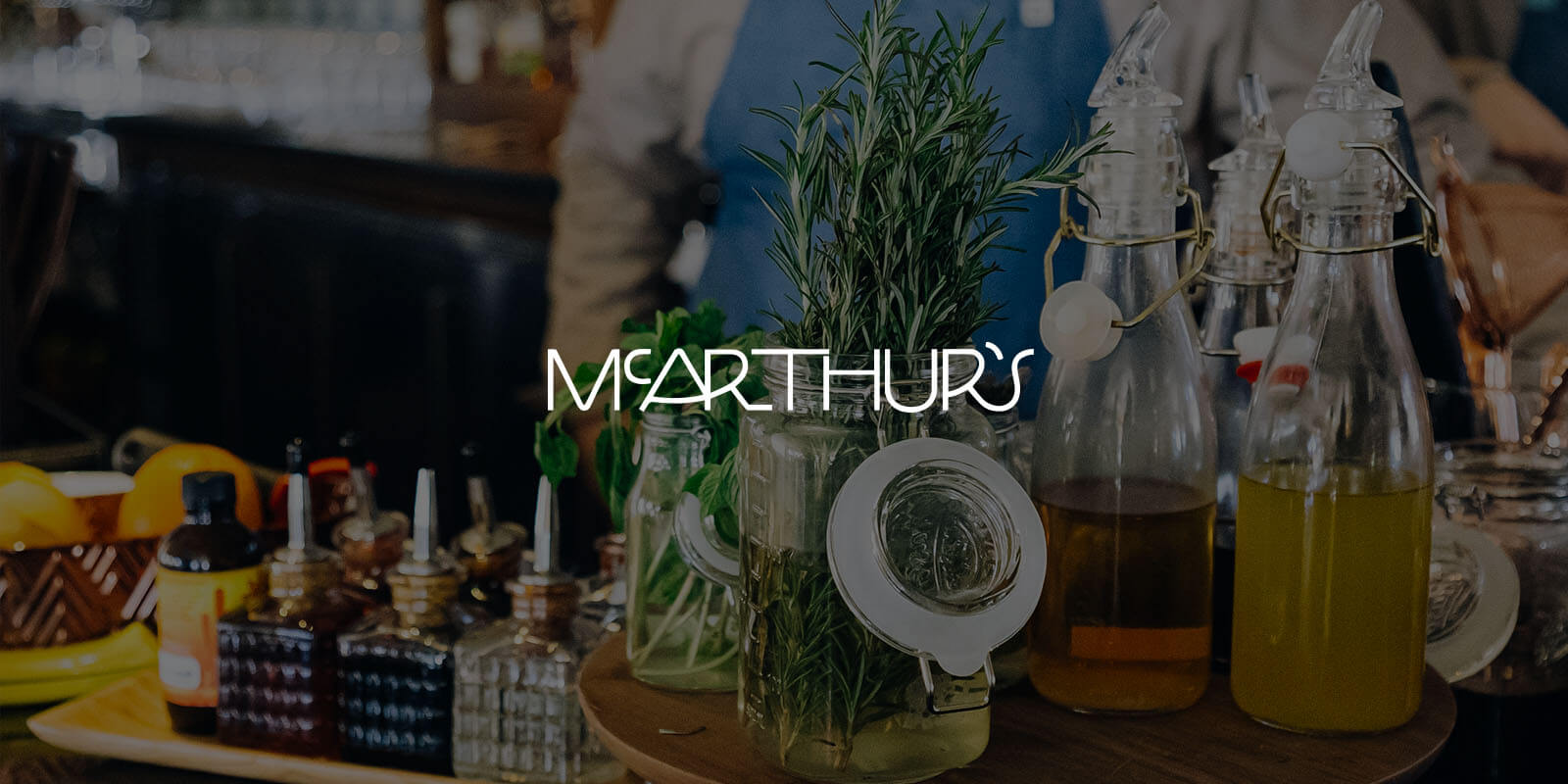 An assortment of glass bottles and jars containing oils, herbs, and spices is displayed on a wooden tray. The background features a blurred figure in a blue apron, indicating a kitchen or bar setting. The text "McArthur's" is overlaid on the center of the image.