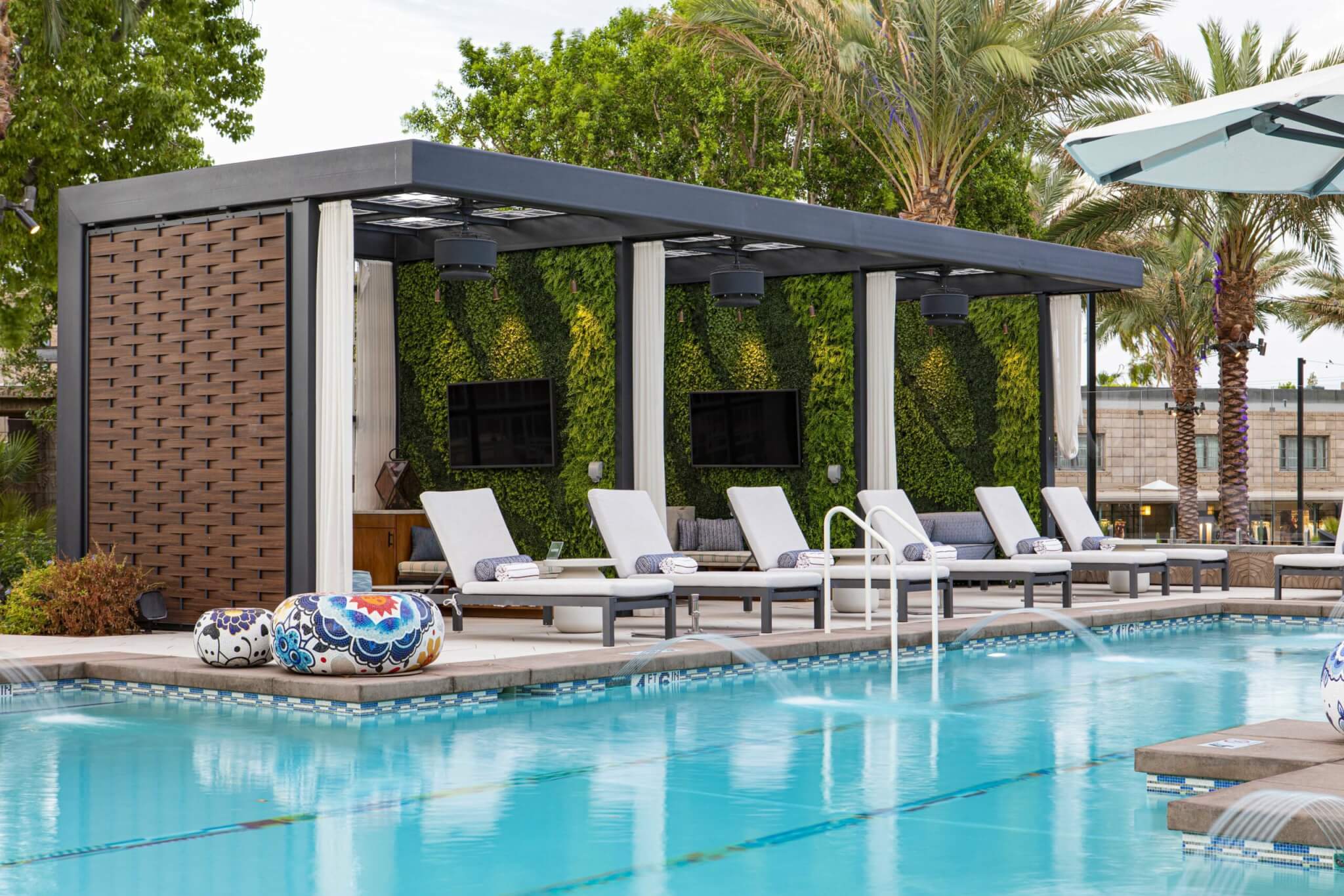 A modern outdoor lounge area beside the Saguaro pool features multiple white poolside chairs under a covered cabana. The cabana has lush green walls, two wall-mounted TVs, and decorative round cushions. The pool includes several water features and palm trees in the background.