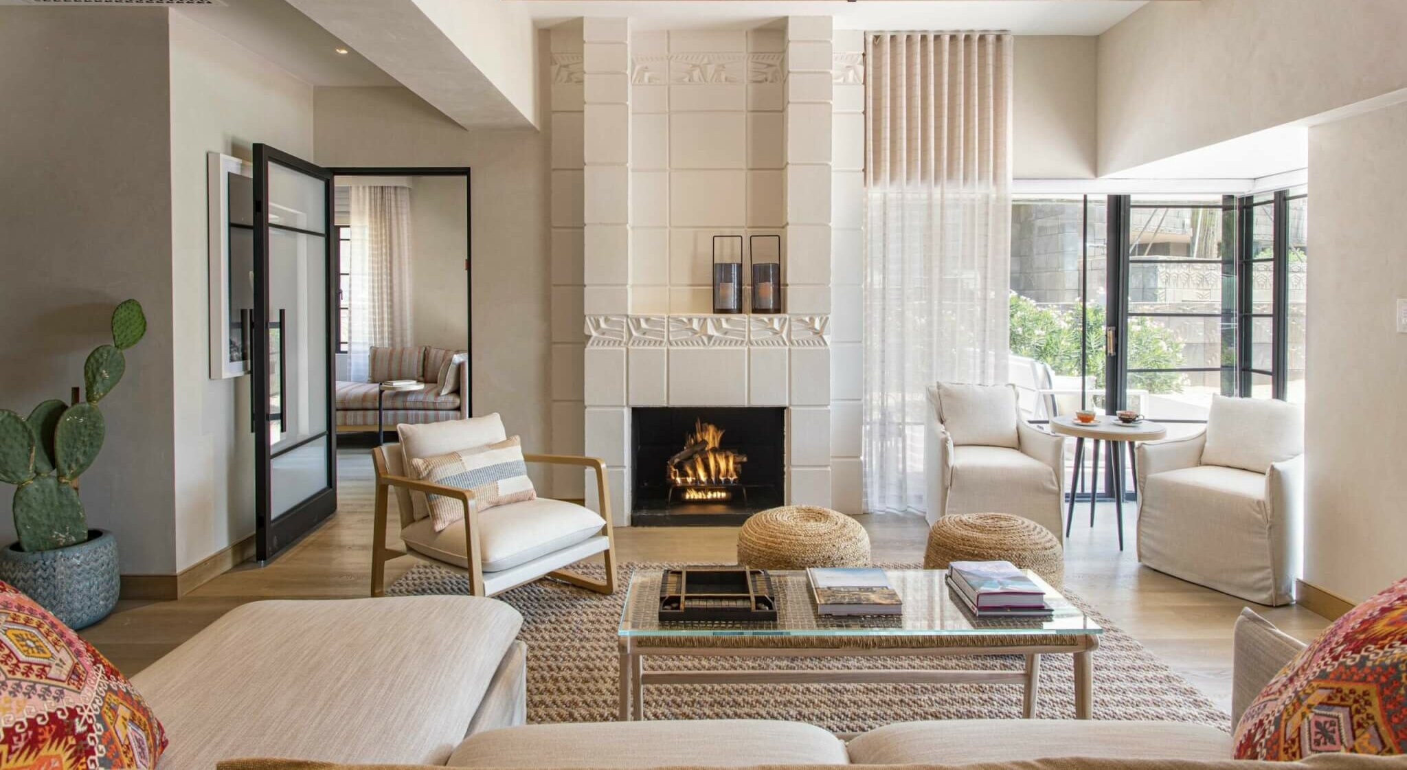 A cozy living room featuring a white fireplace, lit with a warm fire, surrounded by modern furniture. A glass coffee table sits atop a patterned rug, flanked by comfortable chairs and a sofa inviting you to stay. Large windows let in natural light, with a cactus plant in one corner.