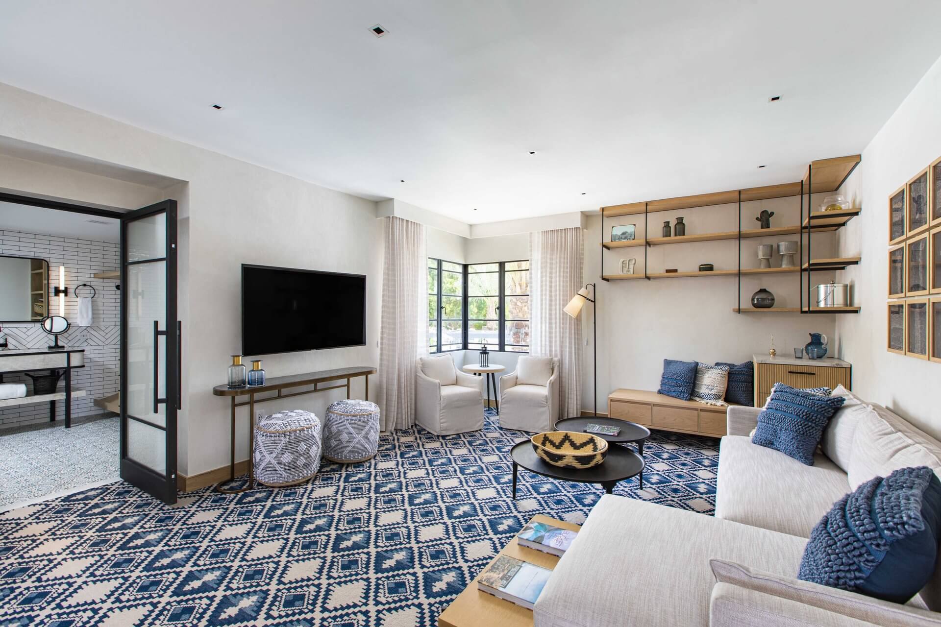 A modern living room featuring a blue and white geometric rug, a white sofa with blue cushions, two white armchairs, and a wall-mounted TV. The room evokes a cozy cottage feel with its large windows, built-in shelves with decor items, and an adjacent bathroom visible through glass-paneled doors.