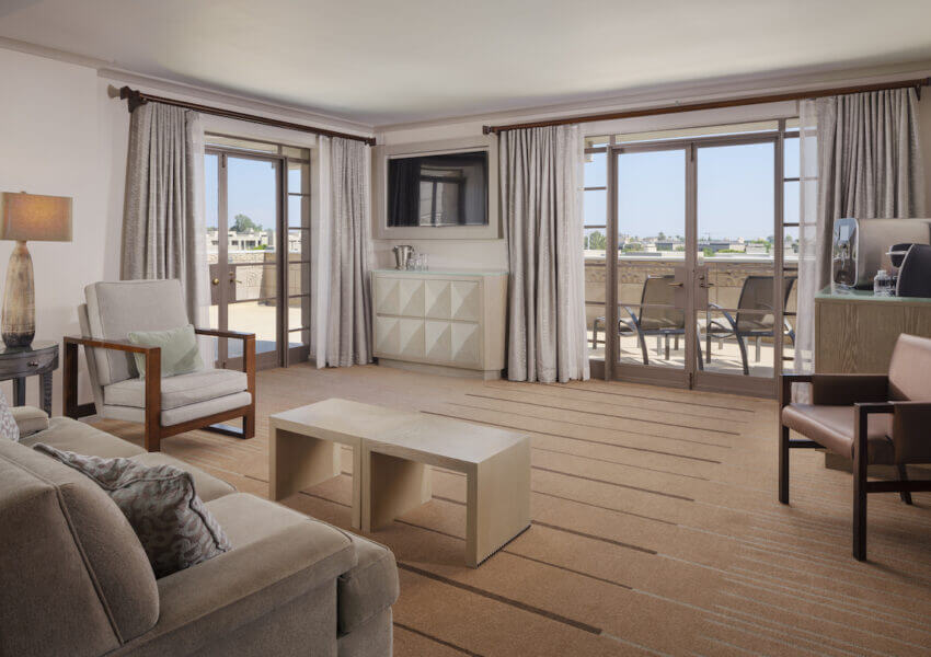 A spacious, modern living room with beige and brown tones. It features a beige couch, wooden coffee table, and two armchairs. Large windows with curtains open to a balcony with outdoor seating. A wall-mounted TV and a sideboard are present. The room is well-lit.