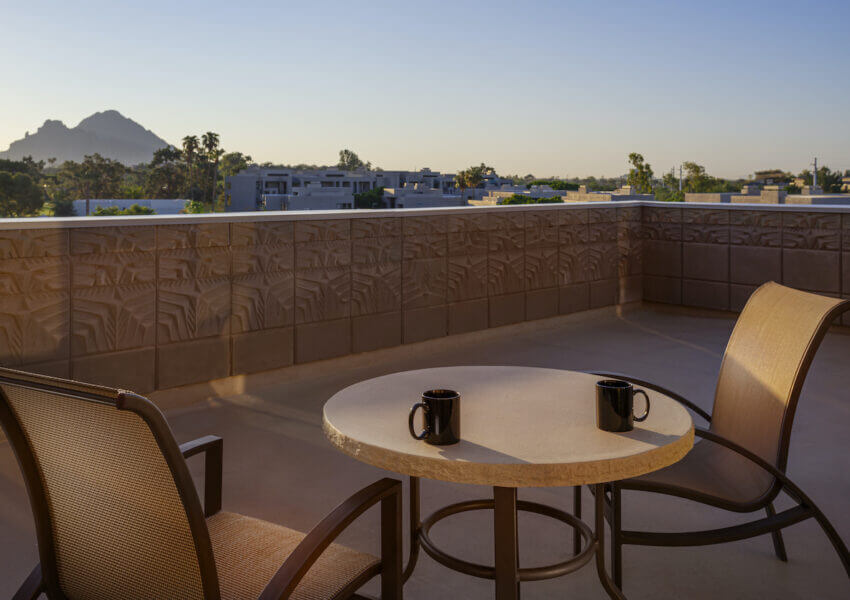 A round outdoor table with two mugs and two chairs on a spacious terrace. In the background, there's a scenic view of distant mountains, lush greenery, and rooftops under a clear sky at dusk.