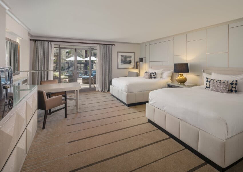 A spacious hotel room with two large beds, each adorned with white linens and decorative pillows. There's a wooden desk and chair in front of a floor-to-ceiling window with sheer curtains, offering a view of the paradise poolside area. Modern, neutral-toned decor completes the room.