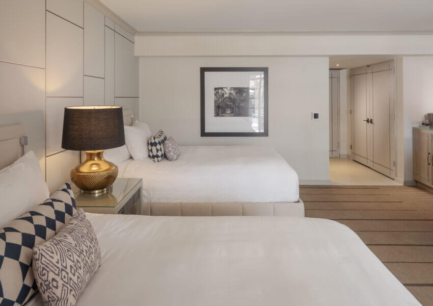 A modern paradise poolside room with two neatly made double beds, each with patterned pillows and a bedside table lamp. The room features minimalistic decor with neutral tones, a framed black-and-white picture, built-in closets, and a small kitchenette area in the background.