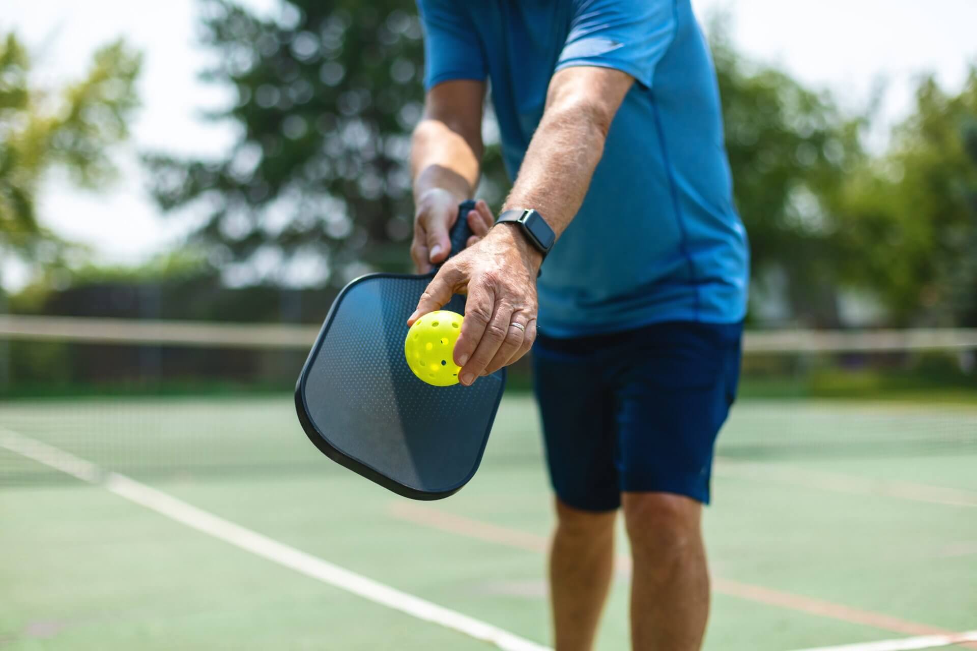 A person wearing a blue shirt and shorts is playing pickleball. They are holding a yellow ball and a black paddle, getting ready to serve on an outdoor court. Green trees and a net are visible in the background.