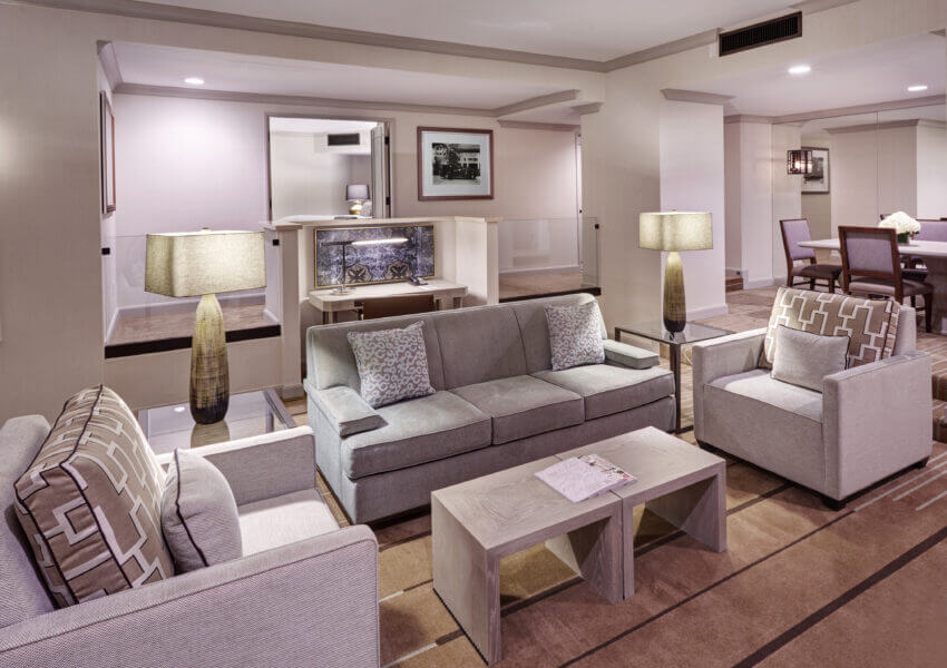 A modern hotel suite featuring a living area with a grey sofa, two armchairs, and a wooden coffee table. Stylish lamps and framed artwork enhance the decor. In the background, there is a dining table with chairs and subtle built-in lighting.