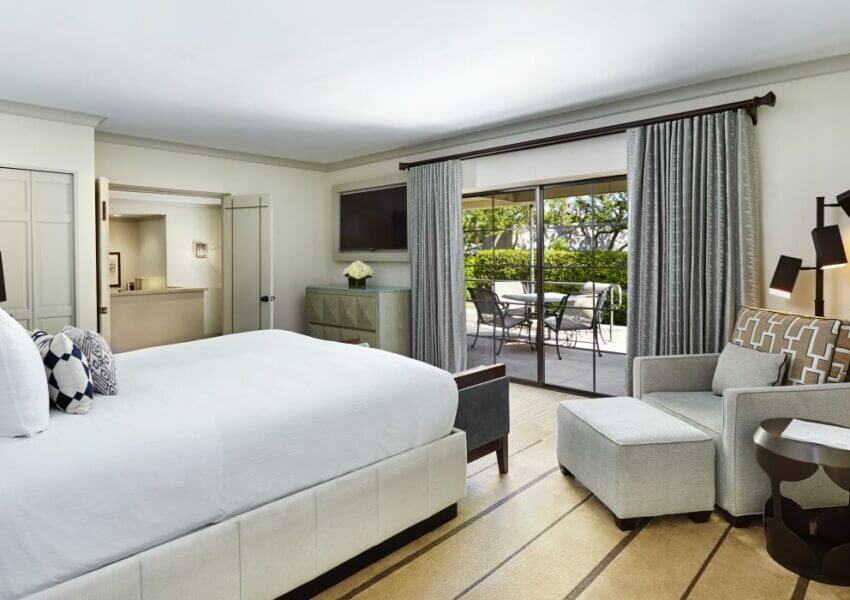 A well-lit signature suite with a large white bed, an armchair with an ottoman, a desk, and a flat-screen TV on the wall. The room has sliding glass doors leading to an outdoor patio with a table and chairs, surrounded by greenery. The decor is modern and elegant.