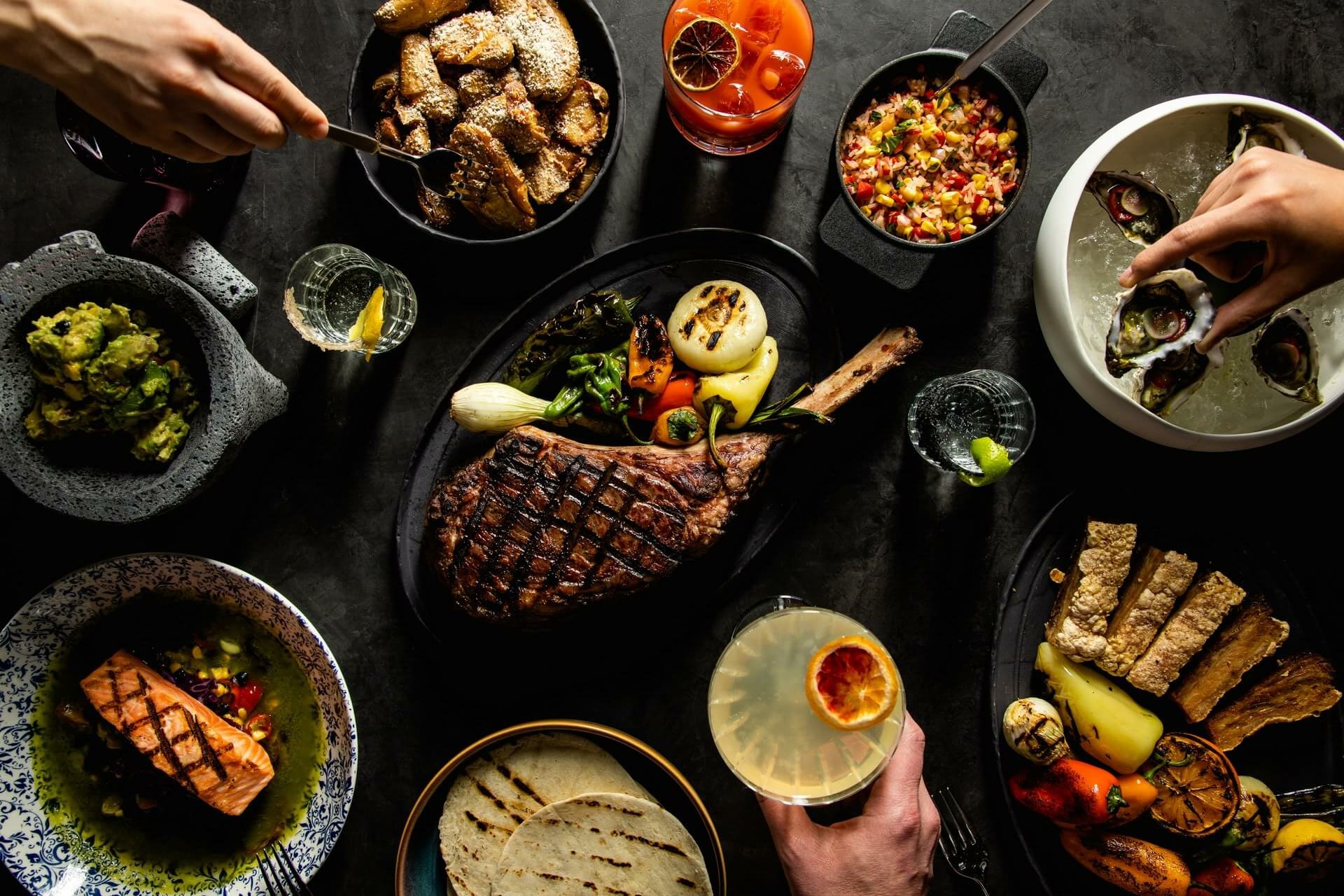 A variety of dishes are laid out on a table, including grilled steak, roasted vegetables, salmon, tacos, dips, and sliced bread. Two hands are seen in the frame—one placing oysters on a platter and another holding a cocktail. Other drinks accompany this spring training feast.