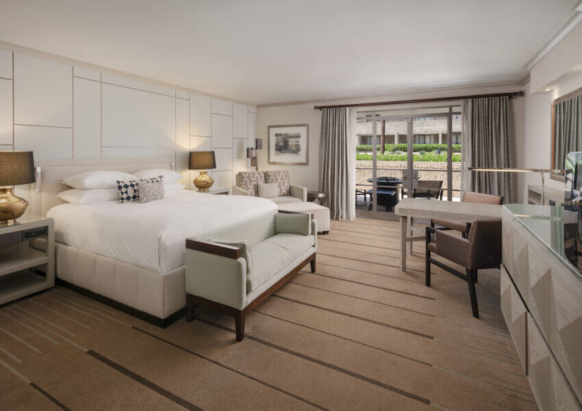 Spacious hotel room with a king-size bed, nightstands with lamps, a bench at the foot of the bed, and a desk with a chair. There is a sitting area with chairs and a table, a large window with curtains, and a view of an outdoor patio and greenery.