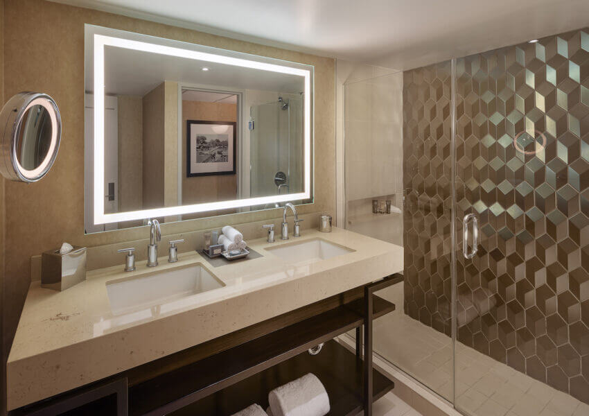 A modern bathroom features a large backlit mirror above a double sink vanity with beige countertops. Towels are neatly placed on a shelf underneath. To the right, a glass-enclosed shower with a geometric tile pattern can be seen. A wall-mounted shaving mirror is on the left.