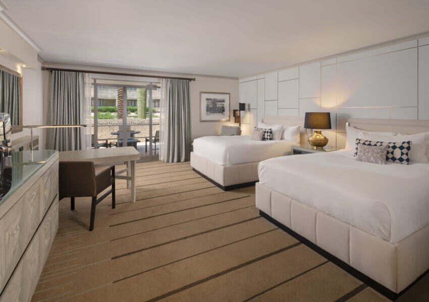 A spacious hotel room with two large beds, a desk and chair, a lamp, and neutral-colored decor. A glass door opens to a balcony with outdoor seating. A flat-screen TV is mounted on a light-colored cabinet along one wall. Walls are decorated with framed art.