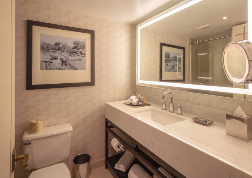 A bathroom with a wall-mounted mirror illuminated by LED lights, a white countertop sink, a toilet with a silver trash can beside it, a rectangular black and white framed photograph on the wall, and beige patterned wallpaper.