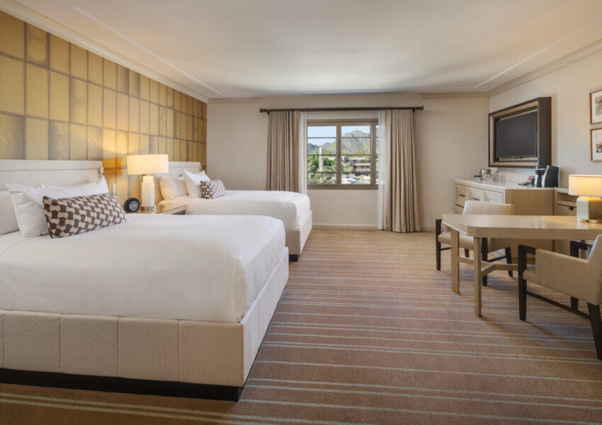 A spacious hotel room with two large white beds, patterned pillows, and a golden accent wall. The room includes a window with a mountain view, a wall-mounted TV, a dresser, a work desk with a chair, a table lamp, and coffee-making facilities.