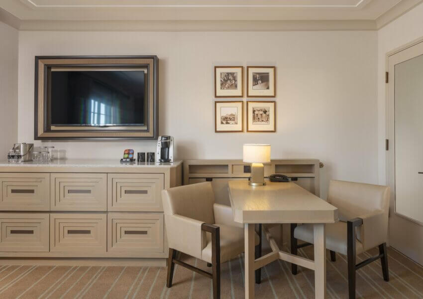 A modern hotel room features a wall-mounted flat-screen TV, a sideboard with a coffee maker and complimentary items, a wooden table with a lamp and phone, two padded chairs, and an accessible shower. Four framed pictures are arranged in a square above the sideboard.